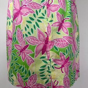 May include: A sleeveless dress featuring a vibrant pattern of pink butterflies and green leaves on a light lime green background. The garment includes a straight hemline with a vertical slit detail positioned at the center of the lower front section.