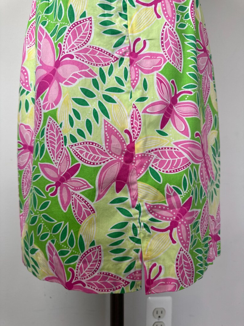 May include: A sleeveless dress featuring a vibrant pattern of pink butterflies and green leaves on a light lime green background. The garment includes a straight hemline with a vertical slit detail positioned at the center of the lower front section.