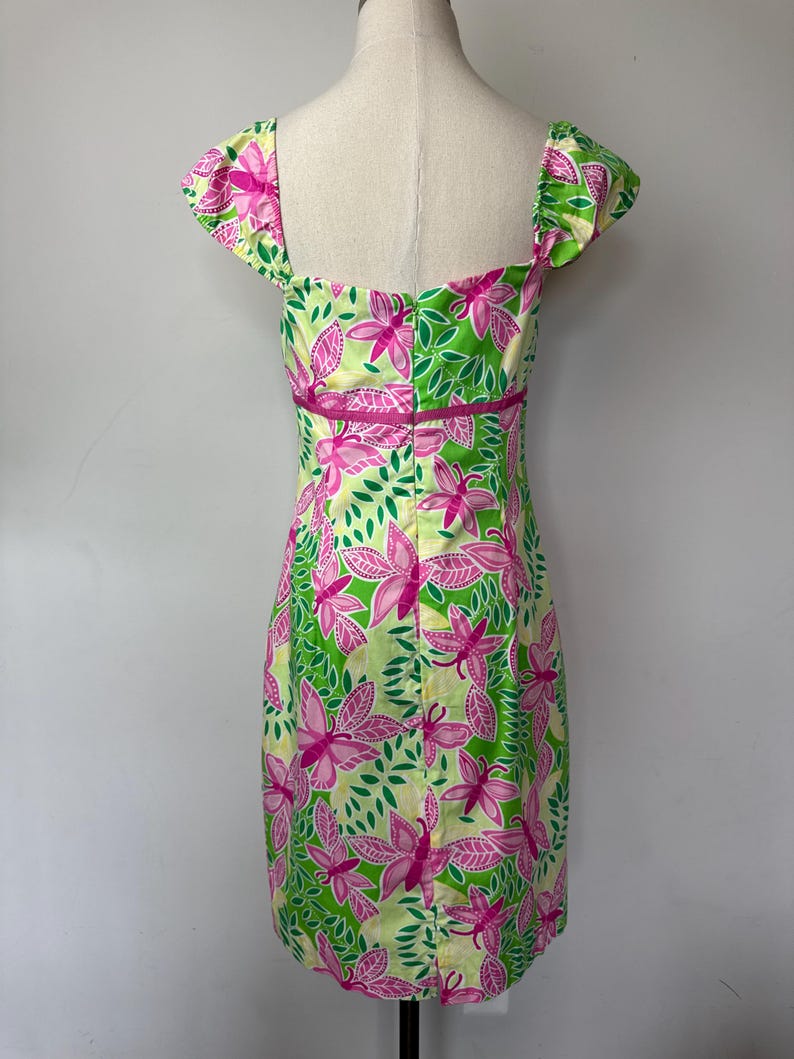 May include: A knee-length dress featuring a vibrant pattern of pink butterflies and green leaves on a pale yellow background. The garment has short cap sleeves, a square neckline, and a horizontal pink band accenting the waistline area.