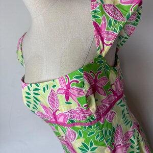 May include: A sleeveless dress featuring a vibrant botanical print with bright pink flowers, lime green leaves, and pale yellow accents. The garment has a square neckline, wide shoulder straps, and a horizontal pink band detail at the waist.