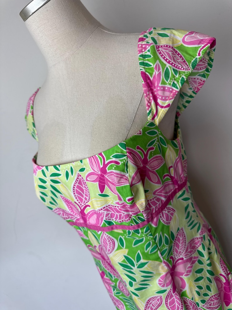 May include: A sleeveless dress featuring a vibrant botanical print with bright pink flowers, lime green leaves, and pale yellow accents. The garment has a square neckline, wide shoulder straps, and a horizontal pink band detail at the waist.