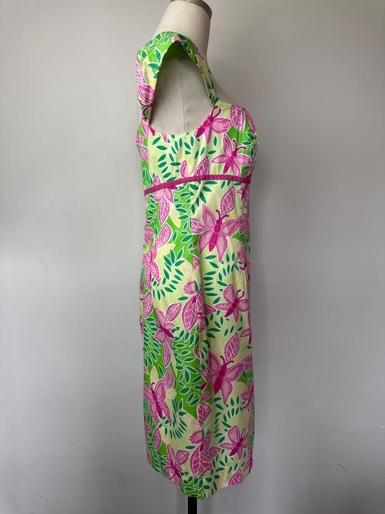 May include: A sleeveless dress featuring a vibrant pattern of pink butterflies and green leaves on a pale yellow background. The dress has a square neckline, a fitted bodice with a horizontal pink band at the waist, and a straight skirt silhouette.