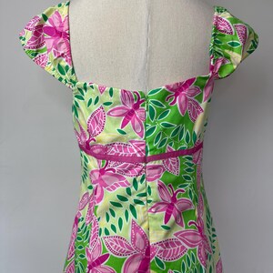 May include: A sleeveless dress featuring a vibrant floral pattern in shades of lime green, bright pink, and yellow. The garment has a square neckline, cap sleeves, and a horizontal pink band at the waist with a central vertical zipper closure.