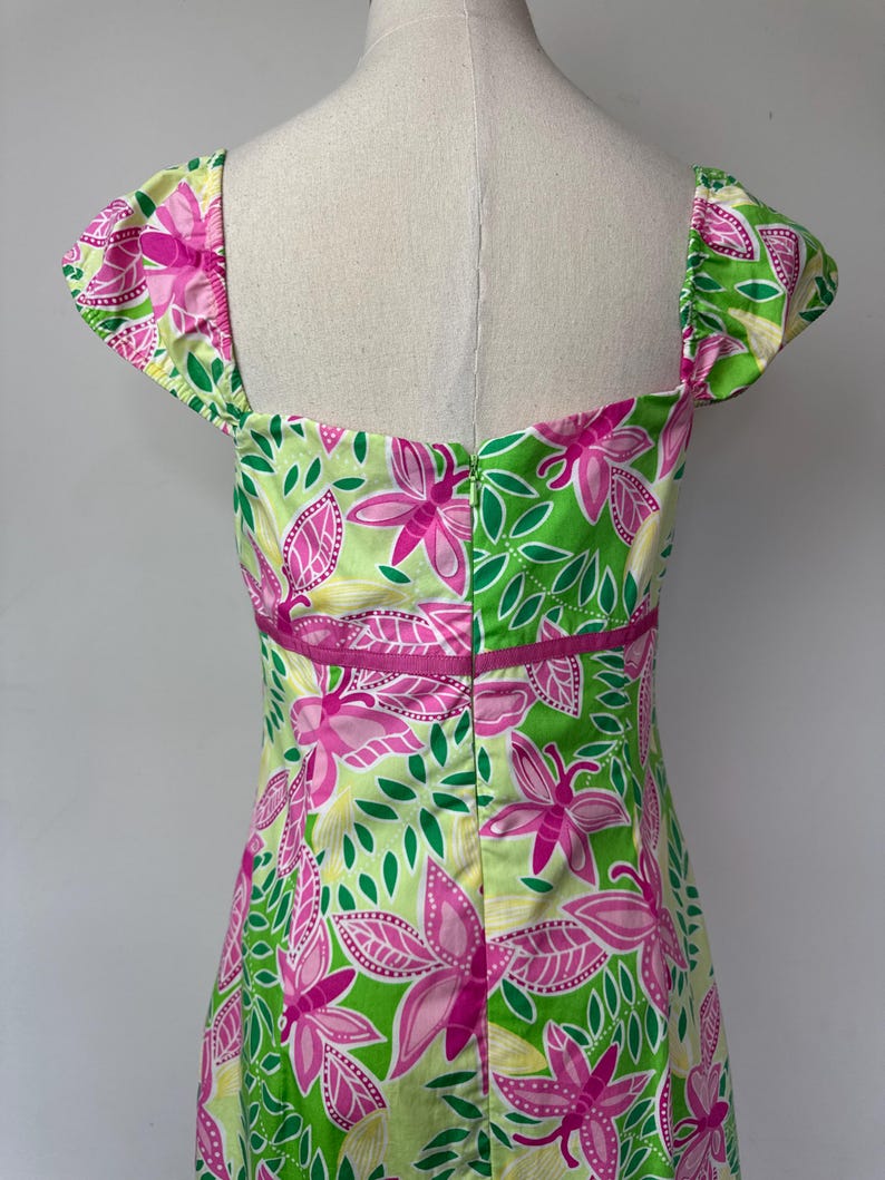 May include: A sleeveless dress featuring a vibrant floral pattern in shades of lime green, bright pink, and yellow. The garment has a square neckline, cap sleeves, and a horizontal pink band at the waist with a central vertical zipper closure.