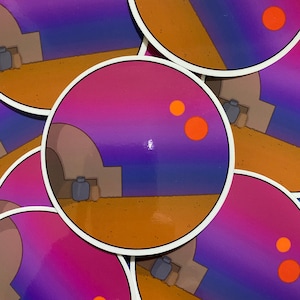 Op de afbeelding: Een sticker met een gestileerde illustratie van een woestijnlandschap met een gebouw op de voorgrond en twee oranje zonnen aan de hemel. De hemel is een gradiënt van paars, roze en blauw.