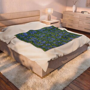 May include: A blue and green patterned throw blanket on a bed. The blanket features a repeating design of blue shapes and yellow lines on a dark green background. The bed has a beige comforter and is in a bedroom setting.