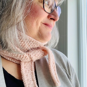 May include: A woman wearing a light pink knitted scarf with a long, thin end that hangs down to her waist. She is wearing a light brown sweater and has gray hair. She is looking out a window.