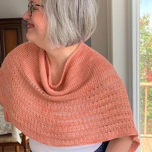 May include: A peach-coloured knitted shawl with a cowl neck. The shawl is draped over the shoulders and has a textured, open-weave design. The person is wearing a white top and blue jeans.