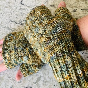 May include: A pair of fingerless gloves knitted with variegated yarn in shades of olive green, mustard yellow, and slate blue. The gloves feature a textured cable knit pattern on the back of the hand and a ribbed cuff that extends up the forearm.