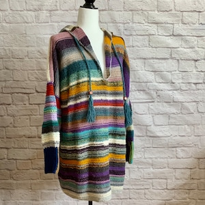 Puede incluir: Un suéter con capucha hecho a mano, multicolor, con cuello en V profundo. El suéter presenta rayas horizontales en varios colores, incluyendo azul, morado, amarillo y rojo. Tiene mangas largas y una capucha con cordones y borlas. El suéter se exhibe en un maniquí.