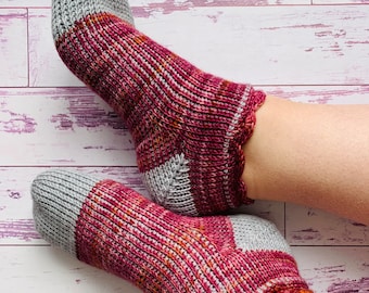 May include: A pair of hand-knitted ankle socks featuring a ribbed texture. The socks are designed with light gray toes and heels, contrasted by a variegated yarn pattern in shades of deep magenta, burgundy, and hints of orange throughout the body.