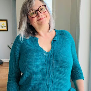 May include: A teal-coloured, long-sleeved, knitted pullover jumper featuring a textured stitch pattern, a split V-neckline, and a vertical decorative panel running down the centre of the chest. The garment has three-quarter length sleeves.