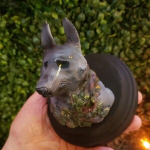 Puede incluir: Escultura gris de la cabeza de un perro con una lágrima, adornada con purpurina dorada y follaje verde. El perro está montado sobre una base negra y circular. La escultura se sostiene en una mano.