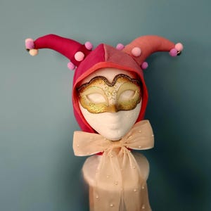 May include: A jester hat in shades of pink and red featuring two long points adorned with small pom-poms and bells. A gold-toned masquerade mask covers the eye area, and a sheer, cream-colored ribbon bow with small pearl accents is tied below.