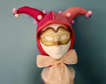 May include: A jester hat in shades of pink and red featuring two points adorned with small pom-poms and bells. A gold-toned masquerade mask covers the eyes of the white mannequin head, and a sheer beige ribbon with pearl accents is tied at the neck.