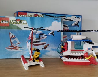 Könnte beinhalten: LEGO System 6342 Rettungshubschrauber-Set mit einem blau-weißen Hubschrauber, einem rot-weißen Rettungsfloß und einem blau-weißen Segelboot. Die Schachtel zeigt den Hubschrauber, der über eine Strandszene fliegt.