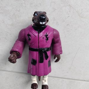 May include: A purple-robed rat action figure with a black belt and a black and white face. The figure has a black and white face with a black nose and a white mouth.