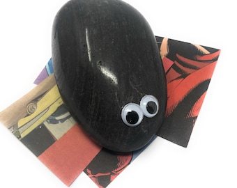 May include: A black rock with googly eyes glued on. The rock is sitting on a colorful comic book page.