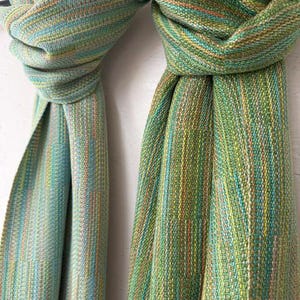May include: A woven scarf featuring a variegated pattern of green, teal, yellow, and orange threads. The fabric displays a textured, multi-coloured striped appearance with a loose, draped knot at the top, showcasing a dense, interlaced weave.