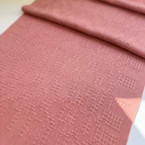 以下が含まれることがあります： 詳細な幾何学模様の、ダスティーローズ色の織物。生地は折りたたまれ、その質感とデザインが明らかになっています。下端にはフリンジが付いています。これは、スカーフ、ランナー、または装飾布である可能性があります。