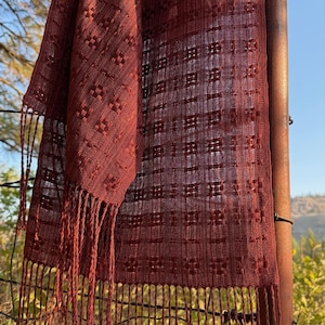 Può includere: Una sciarpa tessuta in una tonalità marrone rossiccio intenso, con un motivo geometrico e bordi sfrangiati. La sciarpa è drappeggiata su una struttura metallica, con uno sfondo naturale all'aperto.
