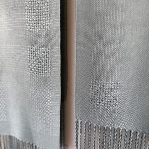 Può includere: Primo piano di due sciarpe azzurre con un motivo quadrato intrecciato. Le sciarpe hanno una frangia sul fondo. Il tessuto ha un aspetto strutturato. Le sciarpe sono appese l'una accanto all'altra.