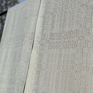 May include: Two panels of light grey fabric with a textured, geometric pattern hanging outdoors. The fabric has a woven appearance with vertical and horizontal lines creating a repeating design. The background shows a blurred view of trees and a blue sky.