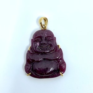 Peut inclure: Un pendentif en pierre sculptée de couleur violette représentant un Bouddha souriant avec des accents dorés.