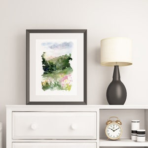 May include: White dresser with three drawers and a shelf. The dresser has a framed watercolor painting of a landscape with green hills and a pink sky. A black lamp with a white shade sits on the dresser. A gold alarm clock and two white candles are on the shelf.