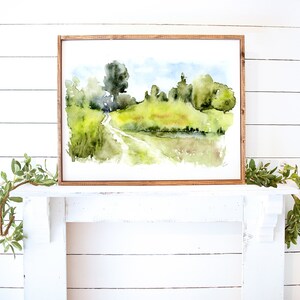 May include: A watercolor painting of a country road winding through a green field with trees and a pond in the background. The painting is framed in a light brown wood frame.