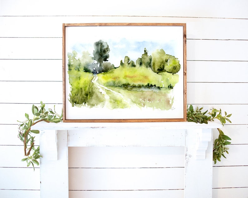 May include: A watercolor painting of a country road winding through a green field with trees and a pond in the background. The painting is framed in a light brown wood frame.