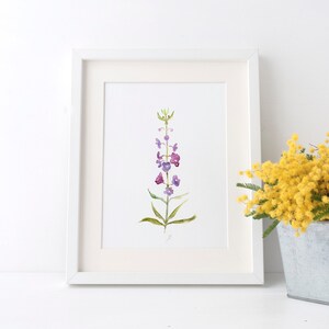 Pode incluir: Uma pintura em aquarela emoldurada de uma flor roxa e verde. A obra de arte est&aacute; em uma moldura branca com uma passe-partout branca. Um buqu&ecirc; de flores amarelas em um recipiente de metal est&aacute; &agrave; direita.