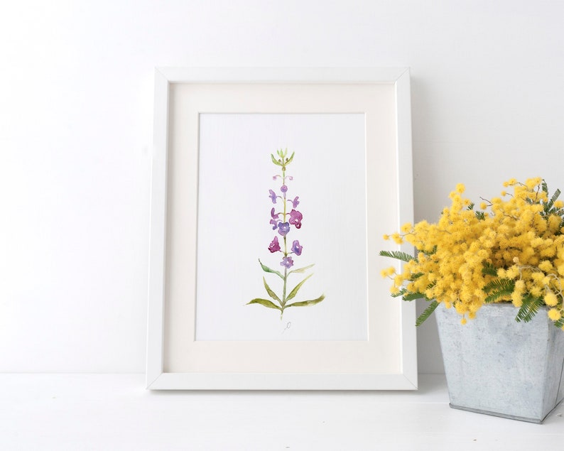 Pode incluir: Uma pintura em aquarela emoldurada de uma flor roxa e verde. A obra de arte est&aacute; em uma moldura branca com uma passe-partout branca. Um buqu&ecirc; de flores amarelas em um recipiente de metal est&aacute; &agrave; direita.