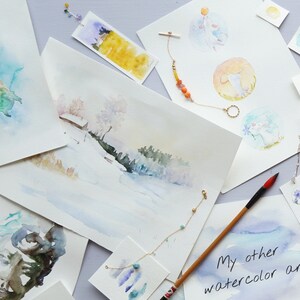 May include: A collection of watercolor paintings on white paper, featuring a variety of subjects including a turtle, a winter scene, and a whimsical animal. The paintings are displayed on a light purple surface with a few decorative items, including a gold necklace with orange beads and a wooden paintbrush.