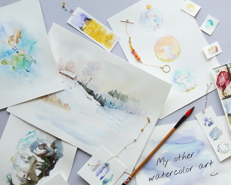 May include: A collection of watercolor paintings on white paper, featuring a variety of subjects including a turtle, a winter scene, and a whimsical animal. The paintings are displayed on a light purple surface with a few decorative items, including a gold necklace with orange beads and a wooden paintbrush.