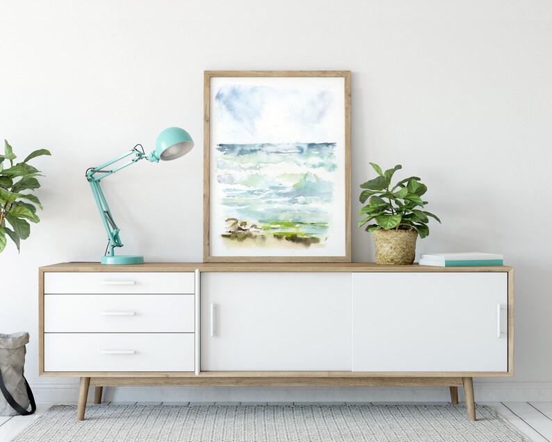 May include: A white dresser with three drawers and two sliding doors. A blue desk lamp is on the left side of the dresser. A potted plant is on the right side of the dresser. A framed watercolor painting of a seascape is above the dresser.