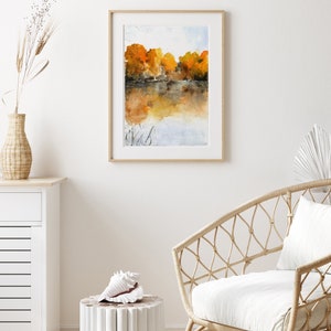 Puede incluir: Una pintura de acuarela enmarcada de un lago con &aacute;rboles en el fondo. Los &aacute;rboles est&aacute;n en tonos de naranja y amarillo, y el agua es de un azul claro. La pintura est&aacute; en un marco de madera simple.
