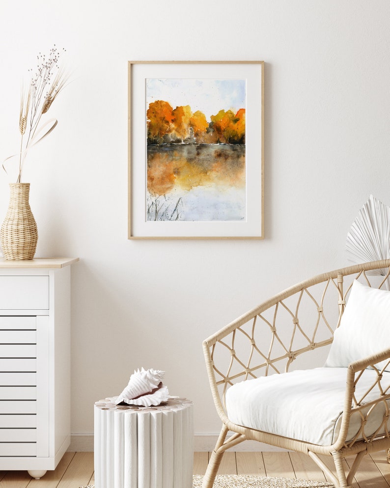 Puede incluir: Una pintura de acuarela enmarcada de un lago con &aacute;rboles en el fondo. Los &aacute;rboles est&aacute;n en tonos de naranja y amarillo, y el agua es de un azul claro. La pintura est&aacute; en un marco de madera simple.