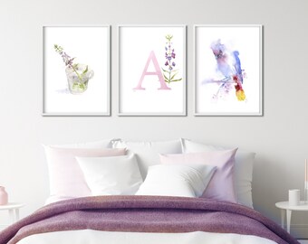 Può includere: Tre stampe ad acquerello che raffigurano una lettera A rosa, un uccello con la coda gialla e un rametto di fiori viola in un bicchiere. Le stampe sono incorniciate di bianco e appese a una parete bianca sopra un letto con una coperta viola.