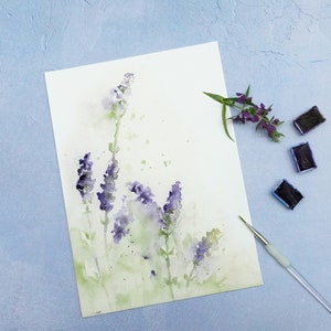 K&ouml;nnte beinhalten: Ein Aquarellbild von Lavendelbl&uuml;ten auf wei&szlig;em Papier. Das Bild ist von Aquarellfarben und einem Pinsel umgeben.