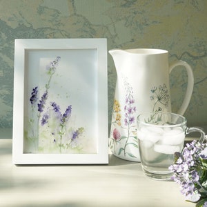 K&ouml;nnte beinhalten: Ein wei&szlig; gerahmtes Aquarellbild von lila Lavendelbl&uuml;ten auf wei&szlig;em Hintergrund. Das Bild steht auf einem Tisch neben einem wei&szlig;en Krug mit Blumendesign und einem Glas Eiswasser.