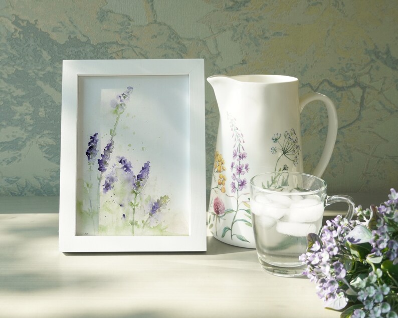 K&ouml;nnte beinhalten: Ein wei&szlig; gerahmtes Aquarellbild von lila Lavendelbl&uuml;ten auf wei&szlig;em Hintergrund. Das Bild steht auf einem Tisch neben einem wei&szlig;en Krug mit Blumendesign und einem Glas Eiswasser.