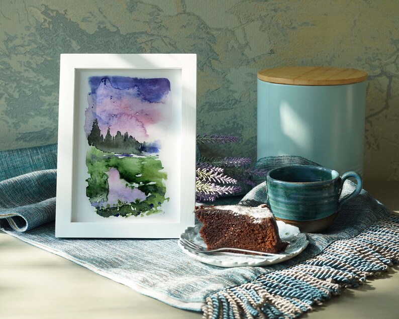 Pode incluir: Uma pintura em aquarela emoldurada retrata uma paisagem com um c&eacute;u roxo e rosa, campos verdes e &aacute;rvores escuras. Uma caneca de cer&acirc;mica azul, uma fatia de bolo de chocolate em um prato pequeno e um recipiente azul com tampa de madeira tamb&eacute;m est&atilde;o presentes.