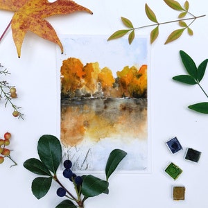 Puede incluir: Pintura acuarela de un paisaje oto&ntilde;al con &aacute;rboles en tonos amarillos y naranjas reflejados en un cuerpo de agua tranquilo. La pintura est&aacute; rodeada de varias hojas y ramas, incluyendo una gran hoja de arce, una rama de bayas y una peque&ntilde;a rama con bayas azules.