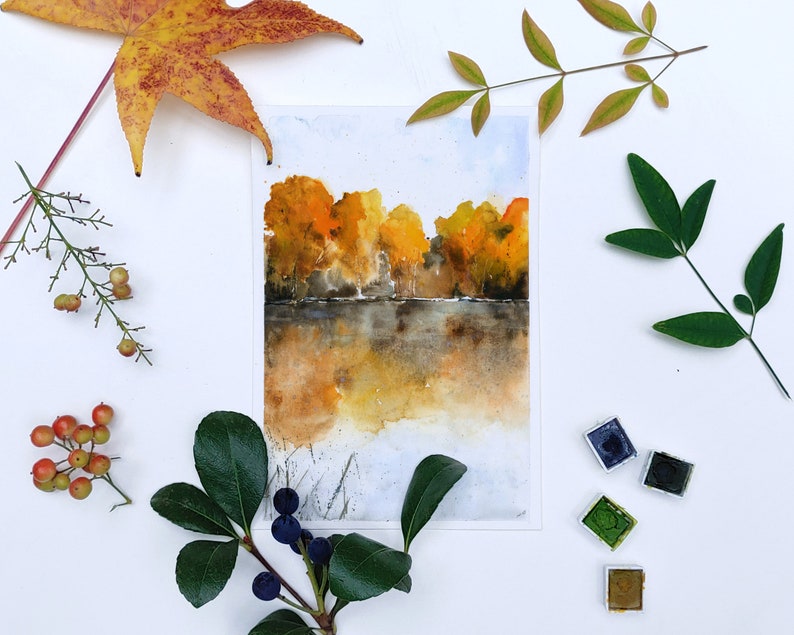 Puede incluir: Pintura acuarela de un paisaje oto&ntilde;al con &aacute;rboles en tonos amarillos y naranjas reflejados en un cuerpo de agua tranquilo. La pintura est&aacute; rodeada de varias hojas y ramas, incluyendo una gran hoja de arce, una rama de bayas y una peque&ntilde;a rama con bayas azules.