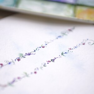 Pode incluir: Um cart&atilde;o branco com um delicado desenho floral em tons de azul, roxo e rosa. O desenho apresenta uma s&eacute;rie de pequenas flores e folhas dispostas em um padr&atilde;o fluido.
