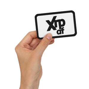 May include: A rectangular patch with a black border and the text "Xrp af" in black letters on a white background. The patch is held by a hand against a white backdrop.