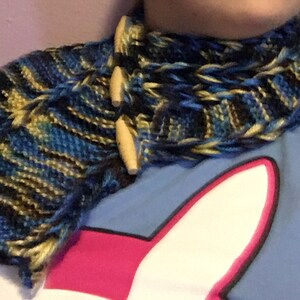 May include: A blue, black, and gold knit scarf with wooden toggles. The scarf is worn around the neck of a person wearing a blue shirt with a pink and white graphic.