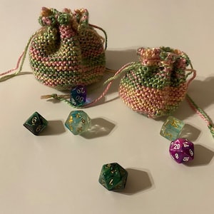 May include: Two crocheted bags in pink and green with a variety of dice, including a green d20, a clear d20, a green d6, a clear d6, and a purple d20.