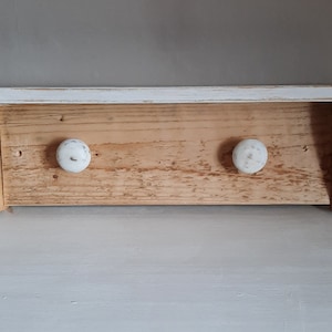 May include: A rustic wooden shelf with a white painted top and two white ceramic knobs. The shelf is made from reclaimed wood and has a distressed finish.